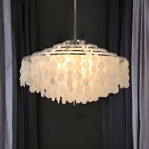 Op de afbeelding: Een witte, gelaagde hanglamp met een zilveren metalen frame. Het licht is versierd met talrijke, ronde, witte schelpachtige elementen, die een zachte, diffuse gloed creëren. De lamp hangt aan een enkel snoer, tegen een achtergrond van grijze gordijnen.