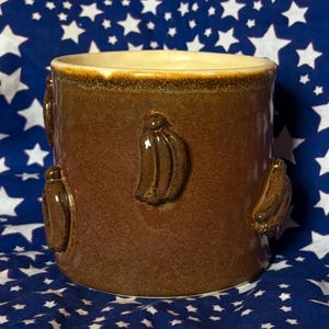 May include: A brown ceramic mug with a glossy finish. The mug features three-dimensional banana-shaped embellishments around its exterior. The interior and rim have a lighter, cream-colored glaze. The mug is cylindrical and appears to be handmade.