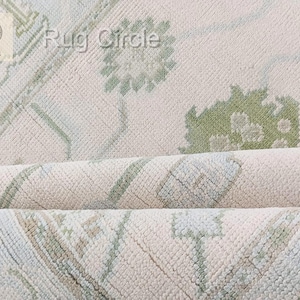 May include: Close-up of a rolled-up, off-white rug with a floral and geometric pattern in shades of green and light blue. The rug has a textured surface and the words "Rug Circle" are visible.