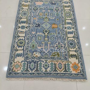 May include: A rectangular, light blue rug with an intricate pattern. The rug features a central design with geometric shapes in shades of blue, green, and orange, framed by a cream-colored border with floral and geometric motifs. The rug has a fringed edge.