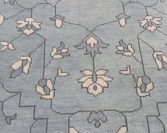 May include: Close-up of a light blue rug with a detailed floral pattern in white and gray. The rug features a repeating design of stylized flowers and geometric shapes, with a fringed edge. The words "Rug Circle" are visible.