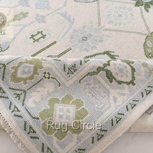 May include: Close-up of a folded area rug with a floral pattern. The rug features a cream-colored background with light blue and green floral designs. The text "Rug Circle" is visible on the rug's surface.