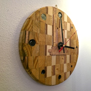 Könnte beinhalten: Eine runde Holzuhr mit einem mosaikartigen Zifferblatt aus verschiedenen Holzstücken. Die Uhr hat schwarze Zeiger, einen roten Sekundenzeiger und schwarze Markierungen. Die Uhr hängt an einer weißen Wand.