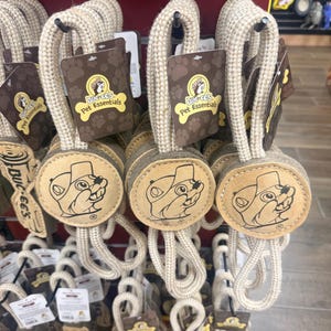 May include: A display of dog toys featuring a round, tan-colored design with a cartoon beaver face and the text "Buc-ee's Pet Essentials." Each toy has a rope handle and a brown tag with the same logo.