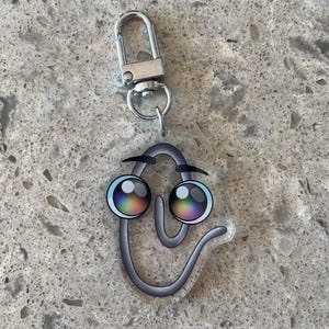 May include: A novelty keychain featuring a smiling paperclip design. The paperclip has large, colorful eyes with rainbow irises and black eyebrows. It is attached to a silver-colored metal clasp.