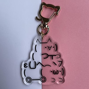 May include: Clear acrylic keychain with a gold-colored cat-shaped clasp. The keychain features a stacked design with black text and emoticons on a white and pink background.