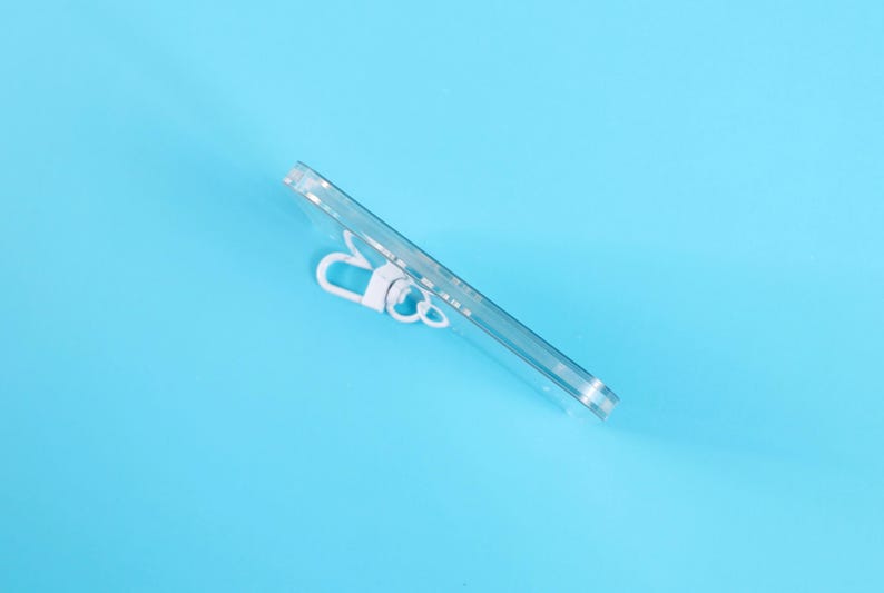 May include: A clear, rectangular phone case with a white clip is angled across a light blue background. The case appears to be made of transparent plastic, offering a view of the phone's back.