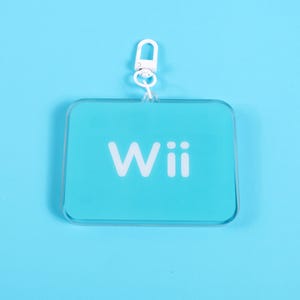 May include: A clear, rectangular keychain with rounded corners and a white metal clasp. The keychain has a light blue background with the word "Wii" printed in white.