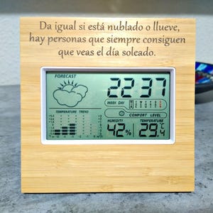 Può includere: Stazione meteorologica digitale con cornice in bambù che mostra ora, temperatura e umidità. Lo schermo mostra un'icona di previsioni, l'andamento della temperatura e il testo "Da igual si está nublado o llueve, hay personas que siempre consiguen que veas el día soleado."