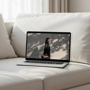 May include: A laptop displaying a fashion advertisement, placed on a white sofa. The laptop screen shows a woman in a black coat holding a handbag, with the text "Albor mock up collection". The sofa is in a well-lit room with a window and curtains.