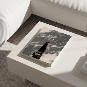 May include: A book titled "Albor mockup collection" rests on a white coffee table. The cover features a woman in a black outfit. The table is in a well-lit room with a white sofa and a light-colored rug.
