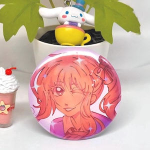 Puede incluir: Un bot&oacute;n redondo rosa con una ilustraci&oacute;n de dibujos animados de una chica gui&ntilde;ando un ojo. La chica tiene el pelo rosa y lleva una camiseta morada. El bot&oacute;n tiene un efecto brillante. Una peque&ntilde;a figura de un personaje de dibujos animados blanco est&aacute; en el fondo.