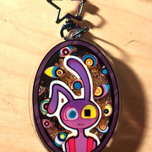 Op de afbeelding: Een ovaalvormige acryl sleutelhanger met een paars konijnfiguur met &eacute;&eacute;n spiraaloog en &eacute;&eacute;n vierkant oog. De achtergrond bevat gouden glitters en meerdere zwevende oogillustraties. Het heeft een stervormige metalen sluiting aan de bovenkant.