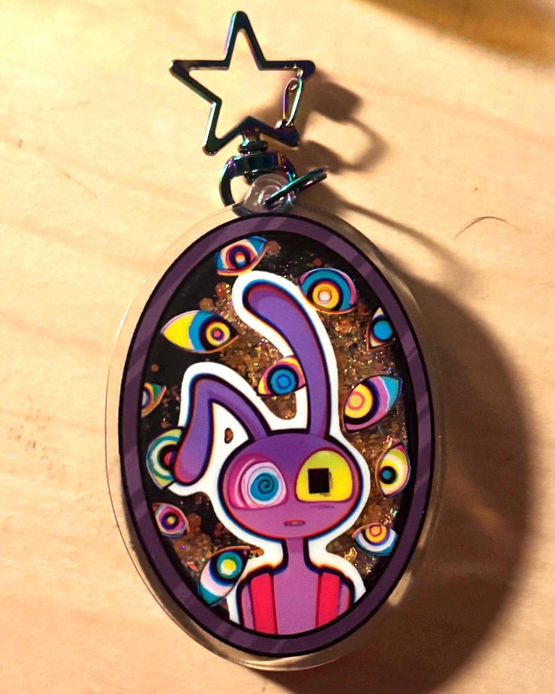 Op de afbeelding: Een ovaalvormige acryl sleutelhanger met een paars konijnfiguur met &eacute;&eacute;n spiraaloog en &eacute;&eacute;n vierkant oog. De achtergrond bevat gouden glitters en meerdere zwevende oogillustraties. Het heeft een stervormige metalen sluiting aan de bovenkant.