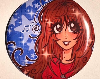 Op de afbeelding: Een ronde button met een anime-stijl illustratie van een persoon met lang bruin haar en grote ogen. De achtergrond is levendig blauw met witte sterren, fonkelingen en muzieknoten. De persoon draagt een rood shirt.
