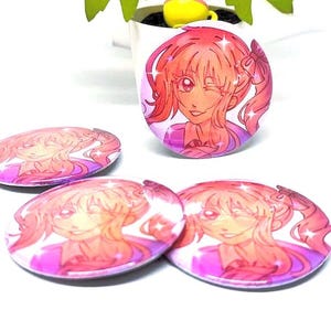 Puede incluir: Varios botones redondos de estilo anime, rosas y morados, con una chica sonriente de pelo rosa y un gui&ntilde;o. Los botones est&aacute;n esparcidos sobre una superficie blanca, con un bot&oacute;n m&aacute;s grande de pie.