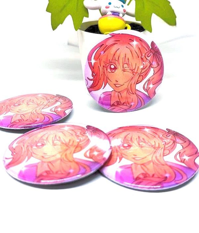 Puede incluir: Varios botones redondos de estilo anime, rosas y morados, con una chica sonriente de pelo rosa y un gui&ntilde;o. Los botones est&aacute;n esparcidos sobre una superficie blanca, con un bot&oacute;n m&aacute;s grande de pie.