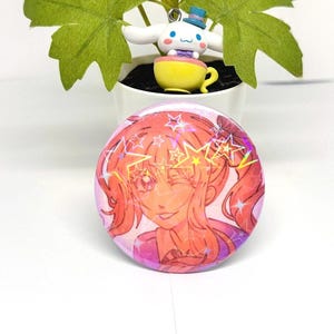 Puede incluir: Un bot&oacute;n redondo rosa con una ilustraci&oacute;n de dibujos animados de una ni&ntilde;a gui&ntilde;ando un ojo, con pelo naranja y un atuendo rosa. El bot&oacute;n tiene detalles de estrellas hologr&aacute;ficas. Una peque&ntilde;a figura de un personaje blanco con un sombrero azul y una taza amarilla est&aacute; en el fondo.