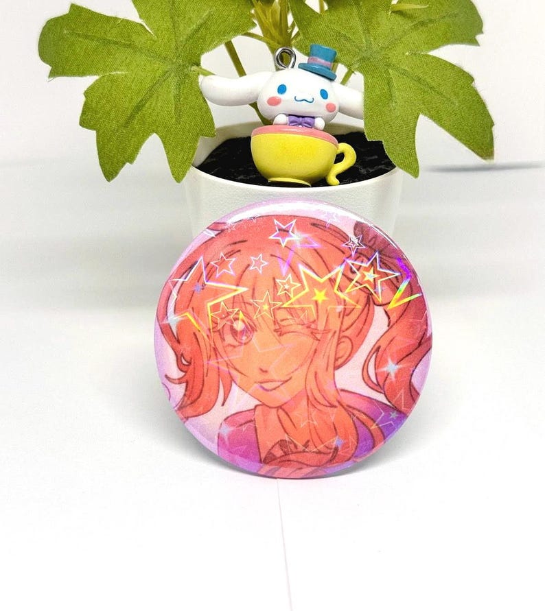 Puede incluir: Un bot&oacute;n redondo rosa con una ilustraci&oacute;n de dibujos animados de una ni&ntilde;a gui&ntilde;ando un ojo, con pelo naranja y un atuendo rosa. El bot&oacute;n tiene detalles de estrellas hologr&aacute;ficas. Una peque&ntilde;a figura de un personaje blanco con un sombrero azul y una taza amarilla est&aacute; en el fondo.