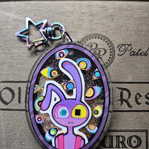 Op de afbeelding: Een ovale acryl sleutelhanger met een paars konijnfiguur met &eacute;&eacute;n spiraaloog en &eacute;&eacute;n vierkant oog, omringd door meerdere zwevende ogen tegen een zwarte glitterachtergrond, bevestigd aan een regenboogkleurige stervormige metalen sluiting.
