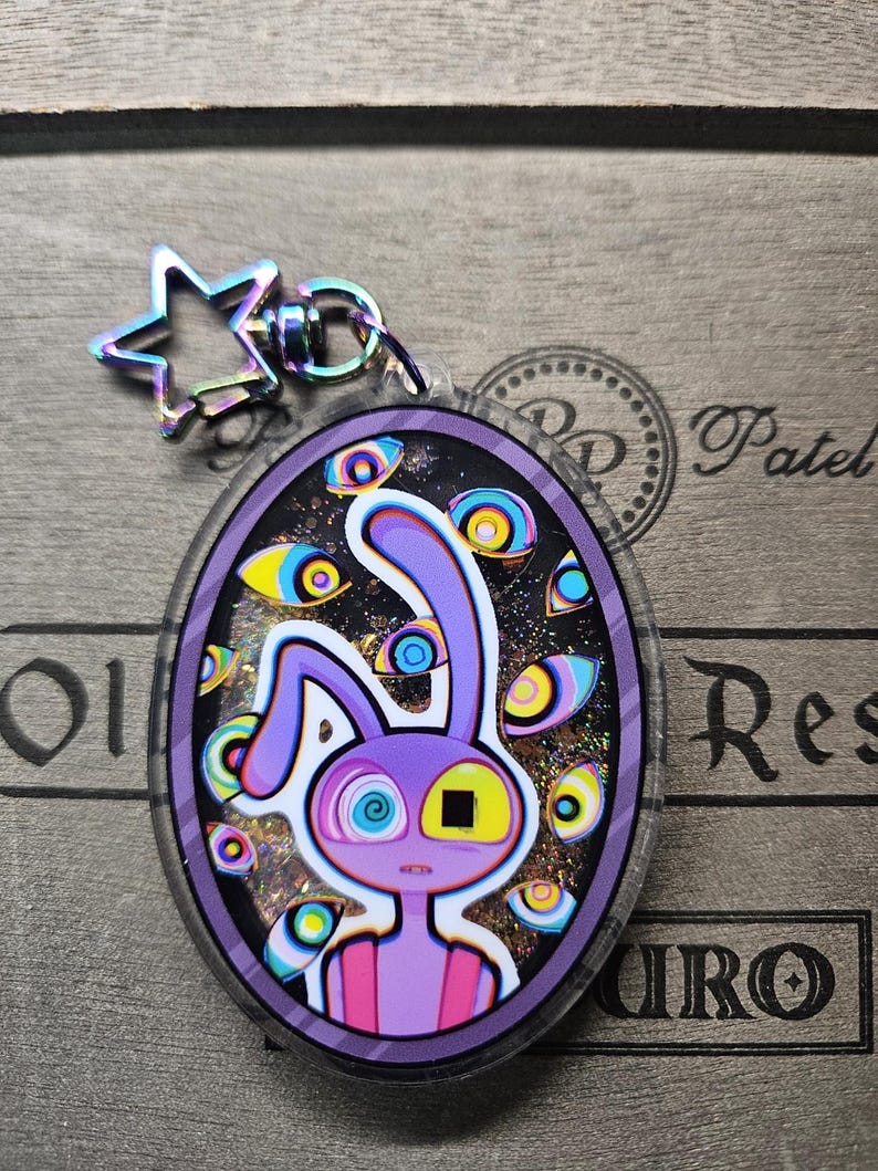 Op de afbeelding: Een ovale acryl sleutelhanger met een paars konijnfiguur met &eacute;&eacute;n spiraaloog en &eacute;&eacute;n vierkant oog, omringd door meerdere zwevende ogen tegen een zwarte glitterachtergrond, bevestigd aan een regenboogkleurige stervormige metalen sluiting.