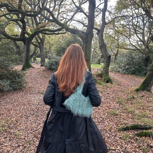 Puede incluir: Un abrigo largo de cuero negro con cinturón a juego y un bolso de mano esponjoso azul claro. La persona camina por un sendero cubierto de hojas caídas, rodeada de árboles con ramas desnudas.