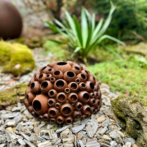 Puede incluir: Una escultura esférica de metal color óxido con numerosas aberturas circulares de varios tamaños. La pieza de arte de jardín se encuentra sobre un lecho de pequeñas piedras grises, rodeada de vegetación y musgo. Otra escultura similar es visible en el fondo.