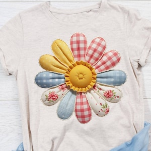 Könnte beinhalten: Ein hellfarbenes T-Shirt mit einer großen aufgenähten Gänseblume. Die Gänseblume hat eine gelbe Mitte und Blütenblätter in verschiedenen Mustern, darunter Vichy, Tupfen und Blumendrucke. Die Blütenblätter sind mit einer weißen Spitzenborte umrandet.