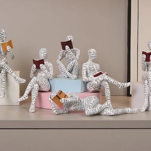 Könnte beinhalten: Eine Sammlung weißer Figuren in verschiedenen Posen, jede mit einem Buch. Die Figuren sind mit Text bedeckt und sitzen auf weißen, rosa und blauen Plattformen. Die Bücher sind braun und rot, was ein literarisches Thema schafft.