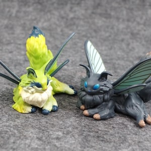 May include: Two fantasy creature figurines. One is lime green with blue accents and a fluffy white chest. The other is dark grey with blue eyes. Both have large, detailed wings and are posed on a grey surface.
