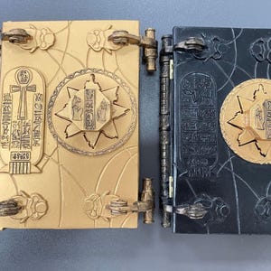 以下が含まれることがあります： 2つの装飾的な本型の箱。1つは金色のヒエログリフと中心のスターバーストデザイン。もう1つは黒色の同様のヒエログリフとスターバースト。どちらも装飾的な留め金とヒンジがあり、古代エジプトをテーマにしているようです。