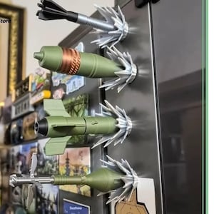 May include: A set of novelty fridge magnets designed to resemble miniature military projectiles. The magnets are in shades of green, black, and silver, with detailed designs. They are attached to a stainless steel fridge door.