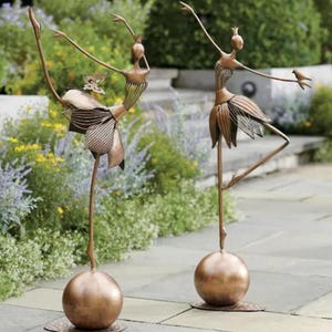 May include: Two bronze metal sculptures of dancing figures. Each figure is balanced on a bronze sphere base. One figure has a butterfly accent, the other has a bird. The sculptures are set in a garden.
