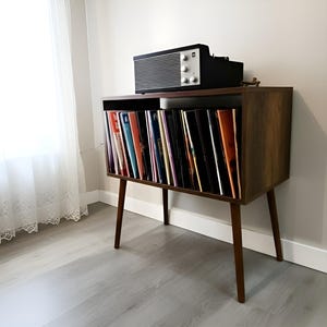 May include: A mid-century modern record player stand with a dark wood finish. The stand features angled legs and a compartment filled with vinyl records. A black and silver stereo sits on top. The room has light gray flooring and a white curtain.