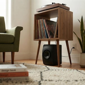 May include: A wooden record player stand with four tapered legs, featuring an open storage compartment filled with vinyl records. A black speaker sits on the floor beneath the stand, and a turntable is positioned on the top surface.