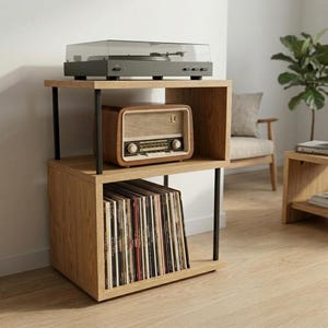 Puede incluir: Un soporte de madera para tocadiscos de dos niveles con acabado en roble claro y soportes de metal negro. El estante superior sostiene un tocadiscos gris oscuro, el estante central una radio de madera y la base almacena discos de vinilo.