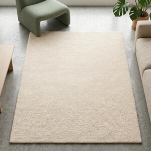 A high-angle view of a plush ivory hand-tufted area rug in a modern living room with concrete flooring, featuring a sage green velvet chair and a minimalist wooden coffee table with pampas grass.