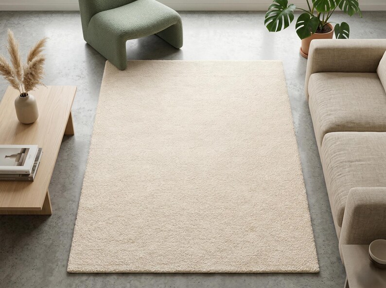 A high-angle view of a plush ivory hand-tufted area rug in a modern living room with concrete flooring, featuring a sage green velvet chair and a minimalist wooden coffee table with pampas grass.