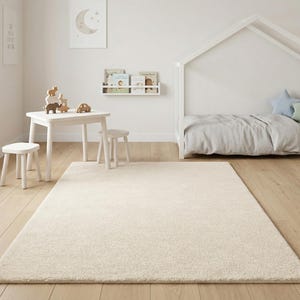 A soft ivory hand-tufted rug in a minimalist Scandinavian-style nursery with a white house-shaped toddler bed, small play table, wooden toys, and light oak flooring.