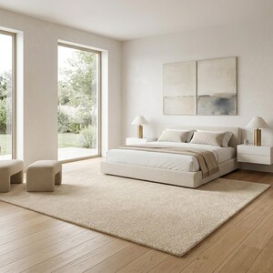 A large beige hand-tufted area rug layered under a modern bed in a sun-drenched minimalist bedroom featuring neutral tones, a cozy accent chair, and light oak wood flooring.