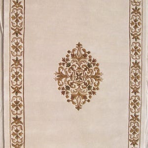 Puede incluir: Alfombra rectangular beige con un borde ornamentado y un medallón central. El borde y el medallón presentan un diseño floral y de volutas detallado en tonos marrón y dorado. El diseño de la alfombra es simétrico y tradicional.