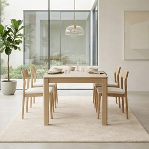 A soft ivory hand-tufted area rug placed under a modern light wood dining table and chairs in a minimalist, sunlit dining room with floor-to-ceiling windows and a large fiddle leaf fig plant.