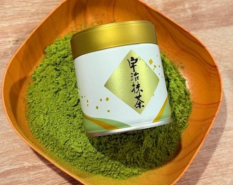 May include: A white and gold matcha tea tin with Japanese text sits in a wooden orange bowl filled with green matcha powder. The tin has a gold lid and a gold diamond-shaped label.