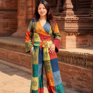 Op de afbeelding: Een patchwork wikkel top en broek met wijde pijpen set. De top heeft lange mouwen en een V-hals. De broek heeft een taille met trekkoord en een patchwork ontwerp met zon- en bloemmotieven. Kleuren: oranje, blauw, groen en rood.