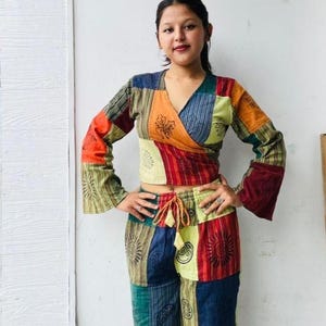 May include: A colourful patchwork two-piece outfit, comprising a wrap top and drawstring trousers. The top has long sleeves with flared cuffs. The outfit is made of fabric squares in blue, orange, red, and green, with sun and paisley designs.