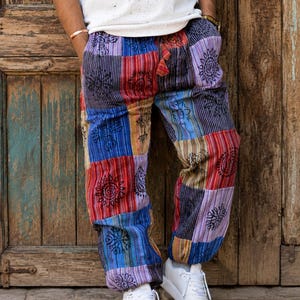 May include: Loose-fitting trousers featuring a patchwork design of various rectangular fabric panels in shades of red, blue, purple, and yellow. The fabric displays black printed circular and floral motifs, with a drawstring waist and elastic cuffs.