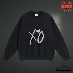 May include: Black sweatshirt with the white text "XO" and a heart graphic. The sweatshirt has long sleeves and a crew neck. A red and white "Best Quality Guaranteed" seal is in the upper right corner. The Azalea Fashion Store logo is at the bottom.