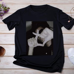 May include: Black t-shirt featuring a sepia-toned image of a person making a hand gesture. The person's hands form a frame, partially obscuring their face. The shirt is made of a smooth, dark fabric.