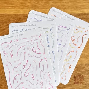 May include: Four sticker sheets featuring arrow designs in various colours. The sheets are labelled "Flow Arrow Stickers" with themes: Berry Gelato, Coastal Tonic, Plum Sorbet, and Caramel Swirl. The arrows are in pinks, blues, purples, and oranges.