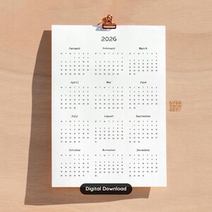 Può includere: Un calendario minimalista del 2026 stampato su carta bianca, tenuto da una clip color rame. I mesi sono disposti in una griglia, con l'anno ben visibile. Le parole "Digital Download" sono in basso.
